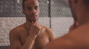 Shirtless man examining himself in the mirror, representing self-reflection and awareness of potential low testosterone symptoms.